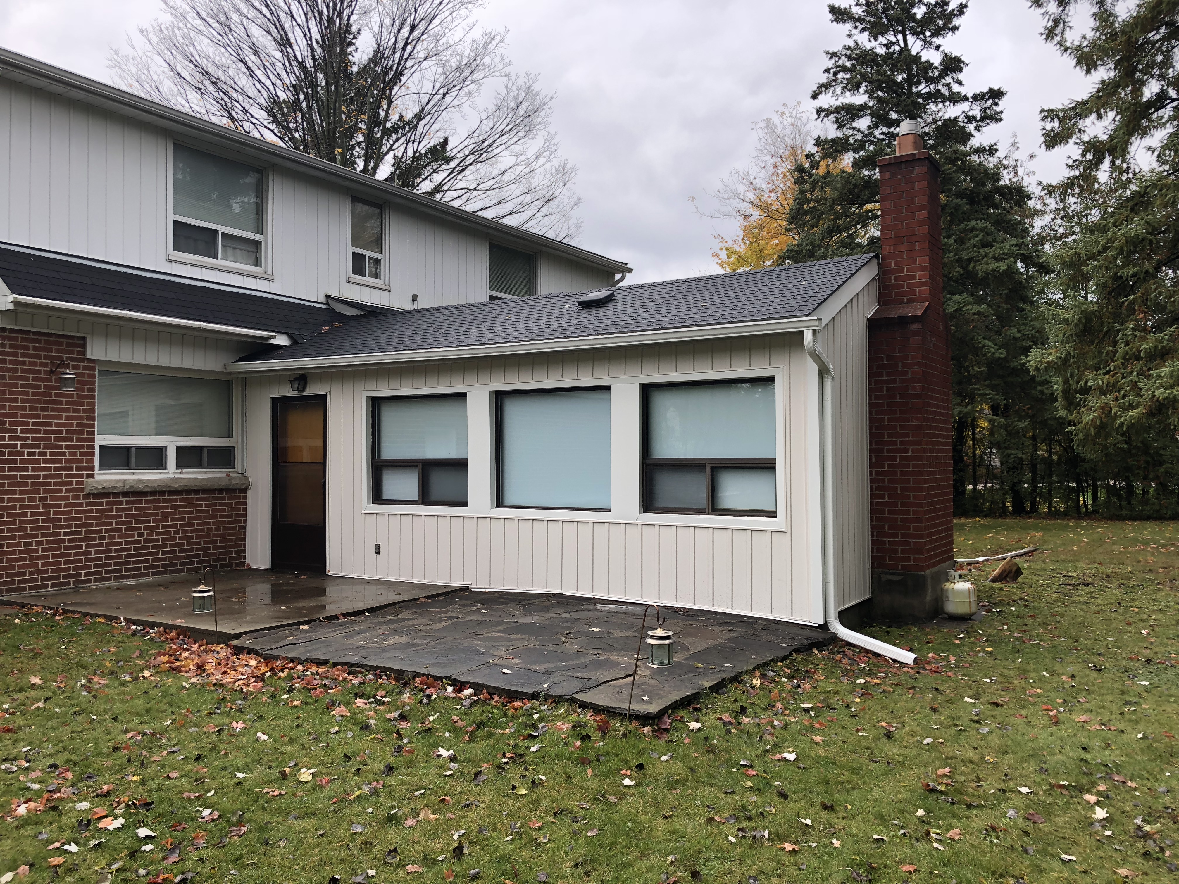 Exterior of a North York home with new vertical siding, updated trim, seamless eavestrough, and a downspout above a stone patio with brick and siding walls.