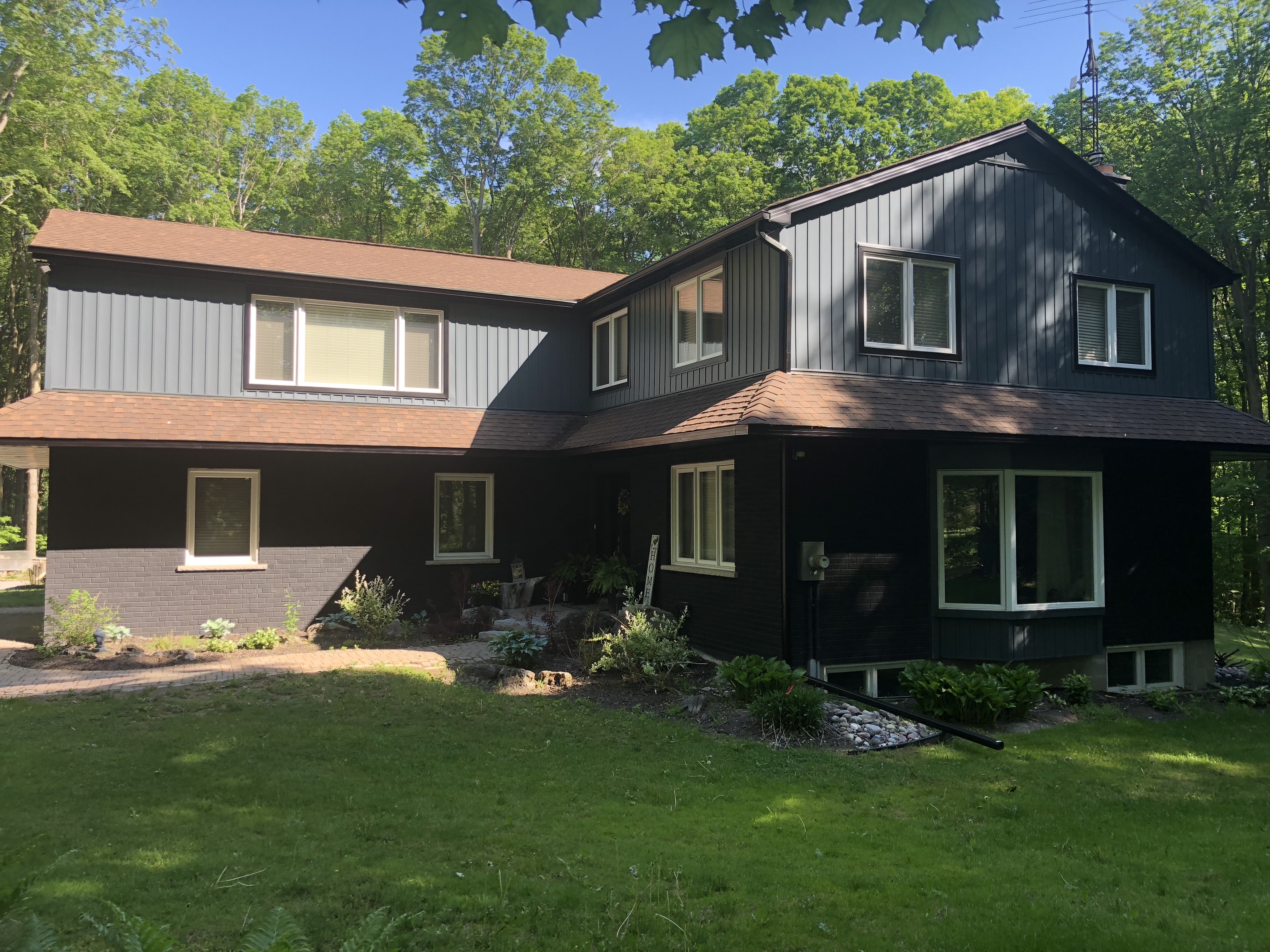 Two‑storey Stouffville home with new dark siding, white trim, landscaped garden beds, and a stone pathway surrounded by tall trees.