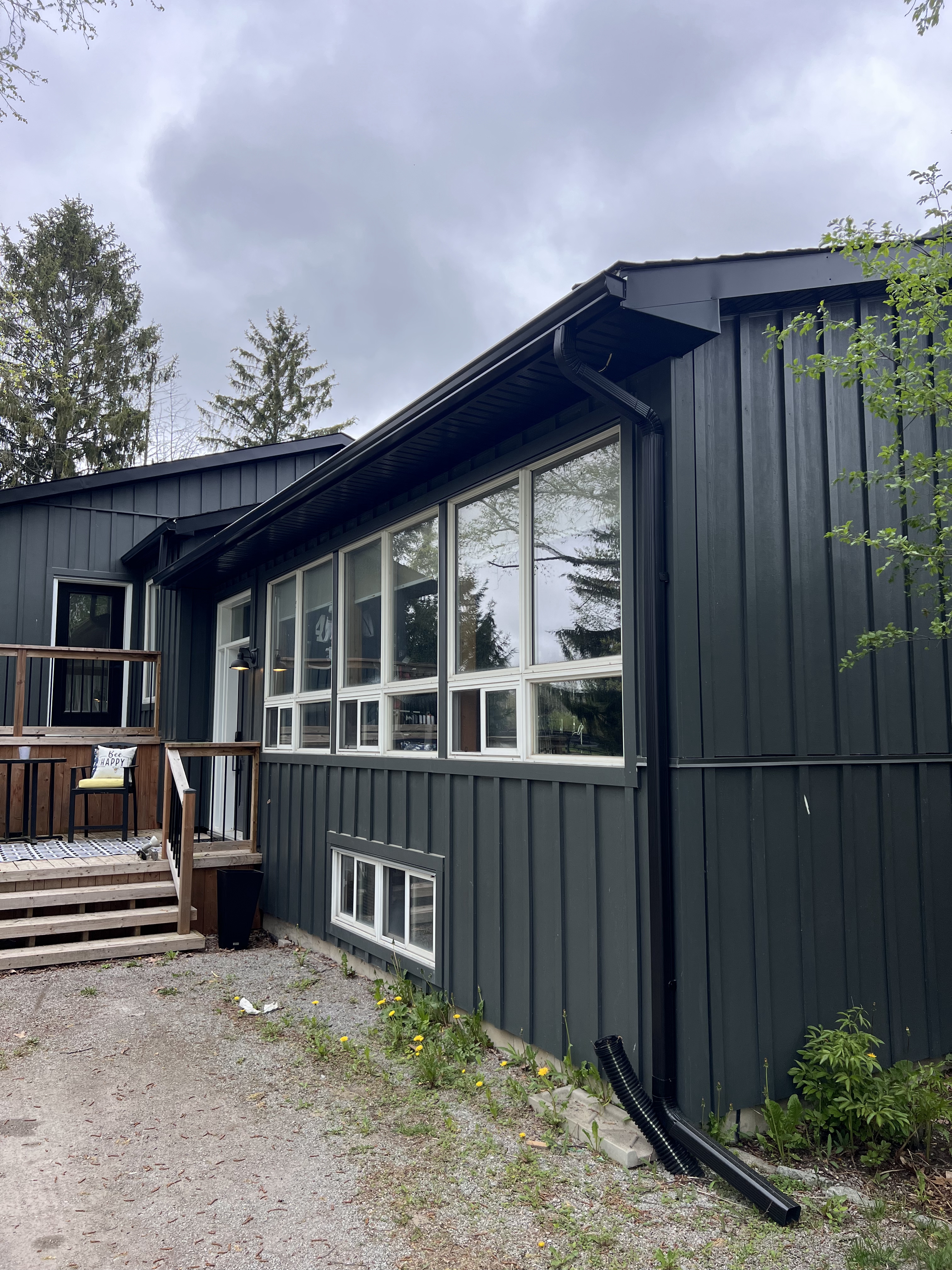 Modern Uxbridge home with new dark vertical siding, white‑trimmed windows, black gutters and downspouts, and a wooden deck surrounded by tall evergreens.