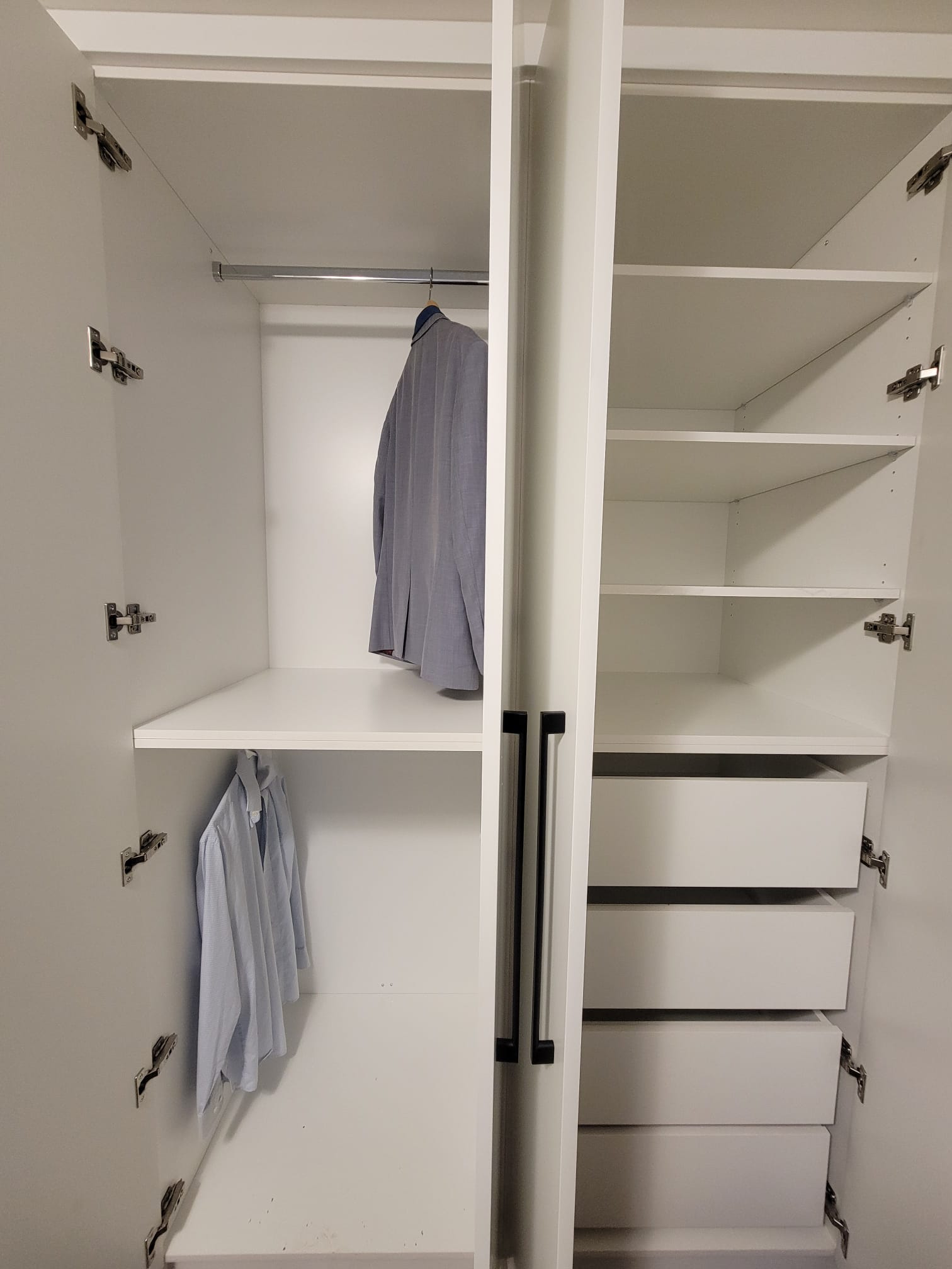 Open white built‑in wardrobe with two hanging sections holding light blue garments on the left and multiple empty shelves and drawers on the right, featuring black handles and a modern organized layout.