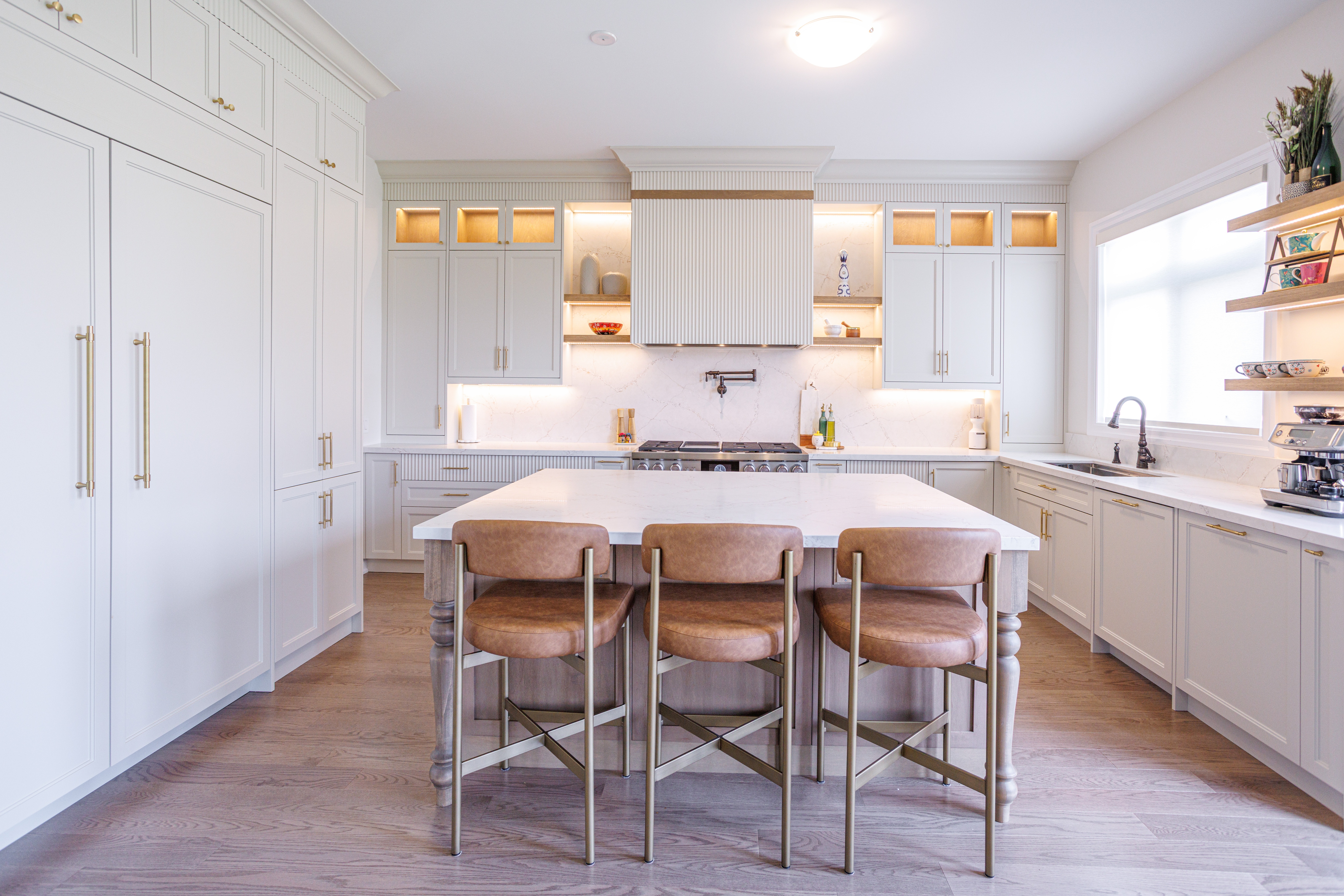 Modern kitchen with a large white marble island, three tan leather barstools with brass frames, light cabinetry with gold hardware, marble backsplash, built‑in stove with pot filler, open shelving, and a sink beneath a large window.