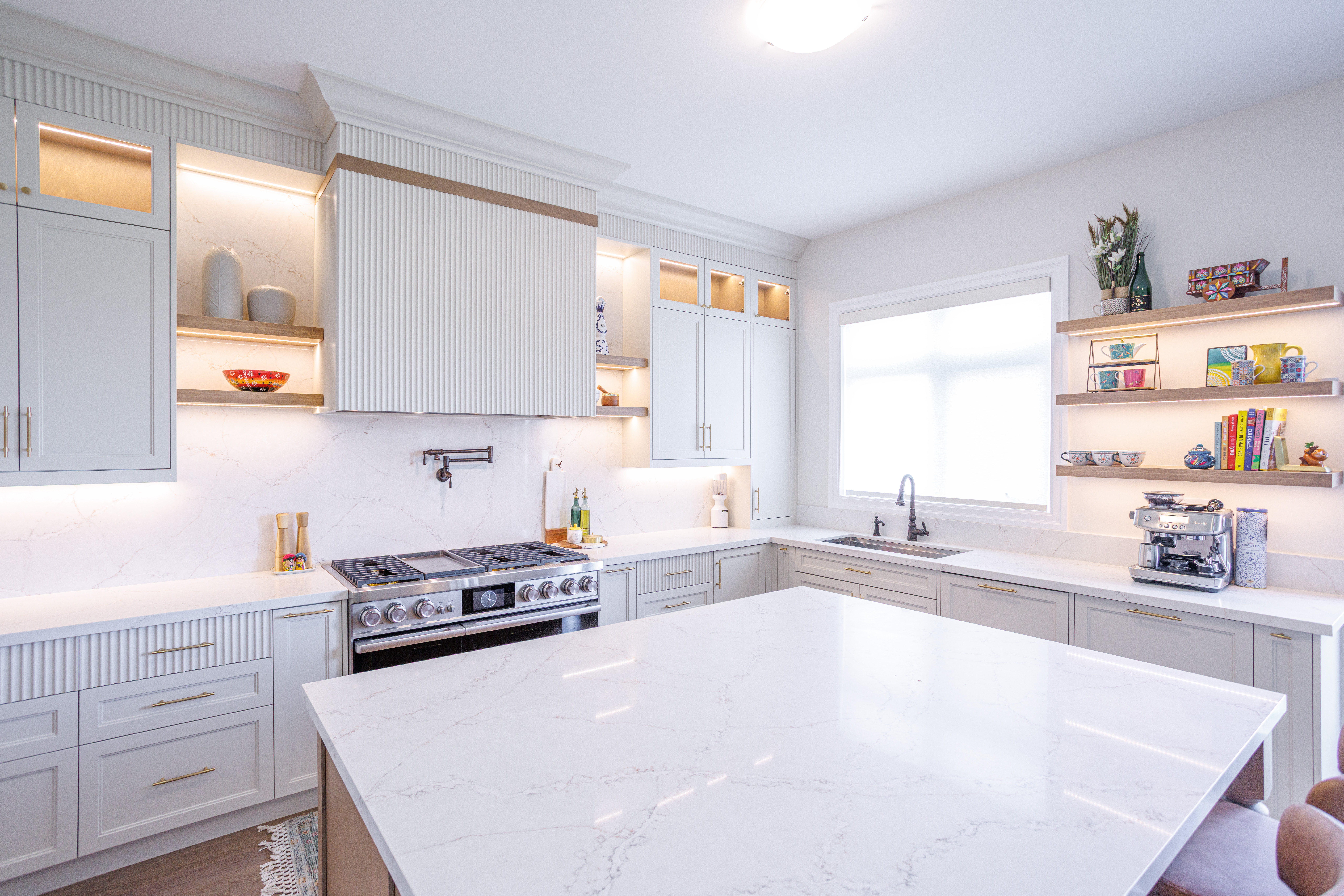Modern kitchen with a large white marble island, light gray cabinetry with gold handles, marble backsplash, stainless steel gas stove with pot filler, open shelves displaying bowls and décor, and a sink beneath a window providing natural light.Modern kitchen with a large white marble island, light gray cabinetry with gold handles, marble backsplash, stainless steel gas stove with pot filler, open shelves displaying bowls and décor, and a sink beneath a window providing natural light.