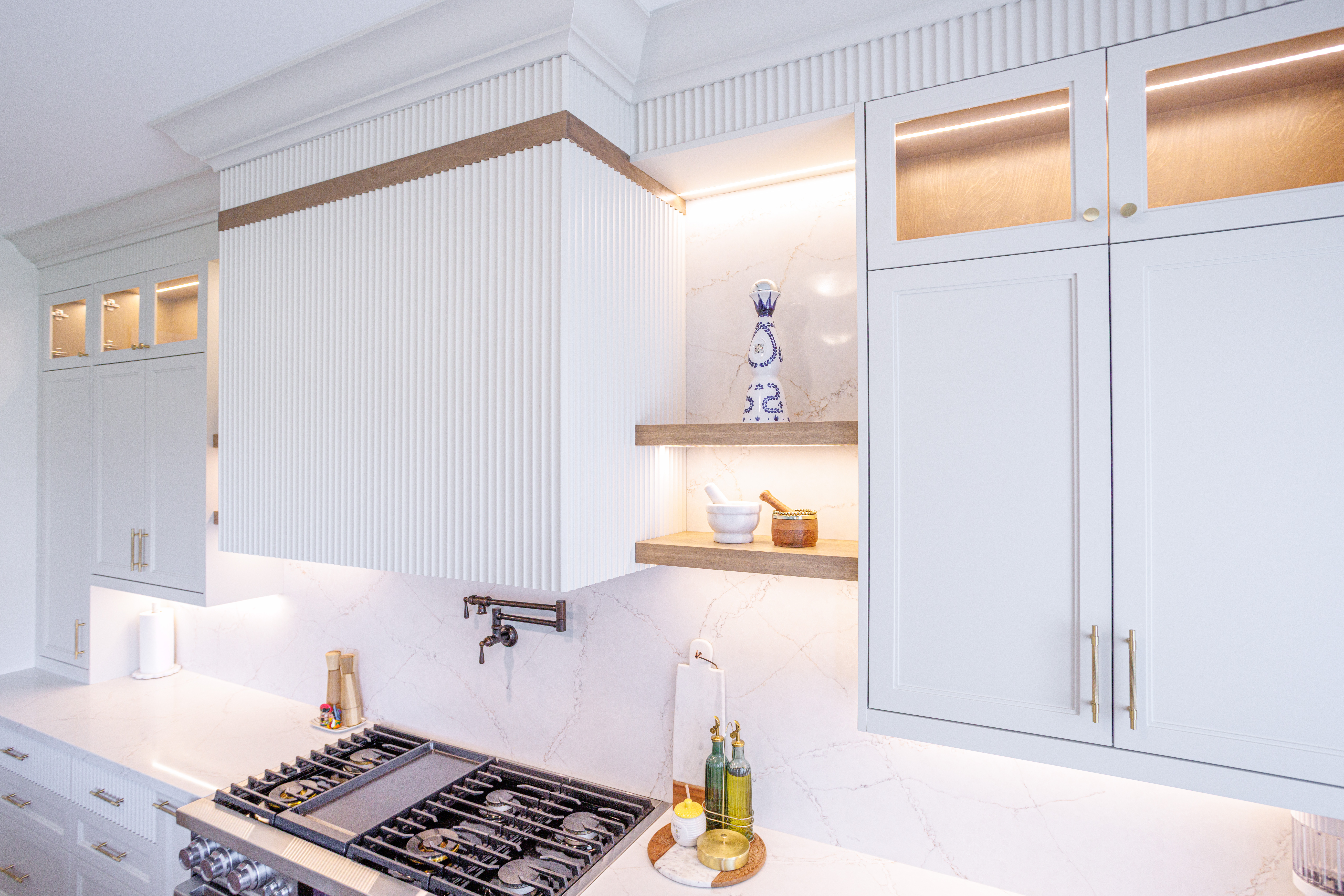 White kitchen cabinetry with gold handles, marble backsplash, stainless steel gas stove with pot filler, fluted upper cabinets with wood trim, and open shelves displaying decorative items and kitchen essentials under warm accent lighting.