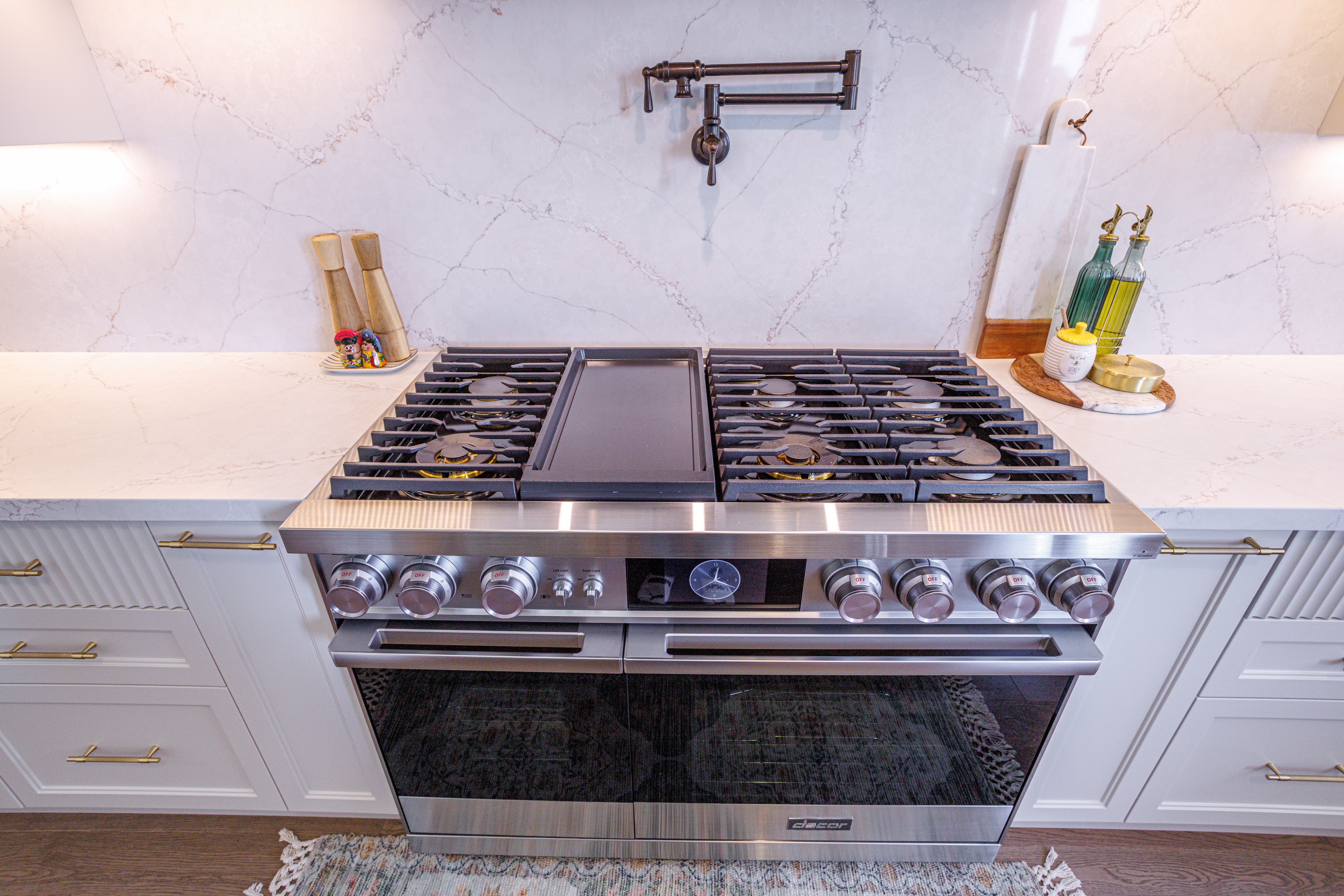Stainless steel gas stove with six burners and a center griddle, set against a marble backsplash with a wall‑mounted pot filler, surrounded by white cabinetry with gold handles and countertop accessories on both sides.
