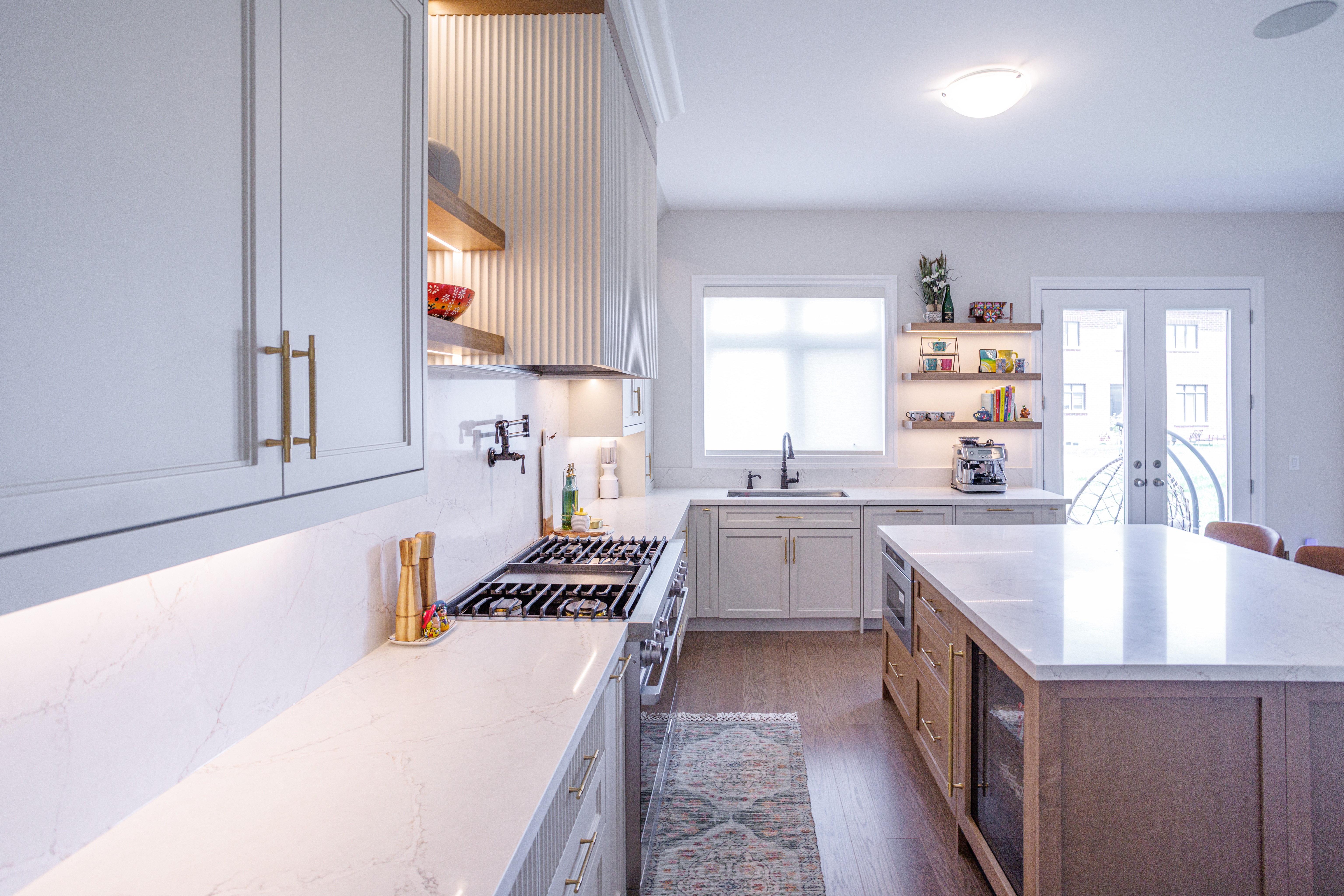 Bright modern kitchen with white cabinetry, gold hardware, marble countertops, wood island base, open shelving, stainless steel appliances, and a patterned runner, all illuminated by natural light from large windows.