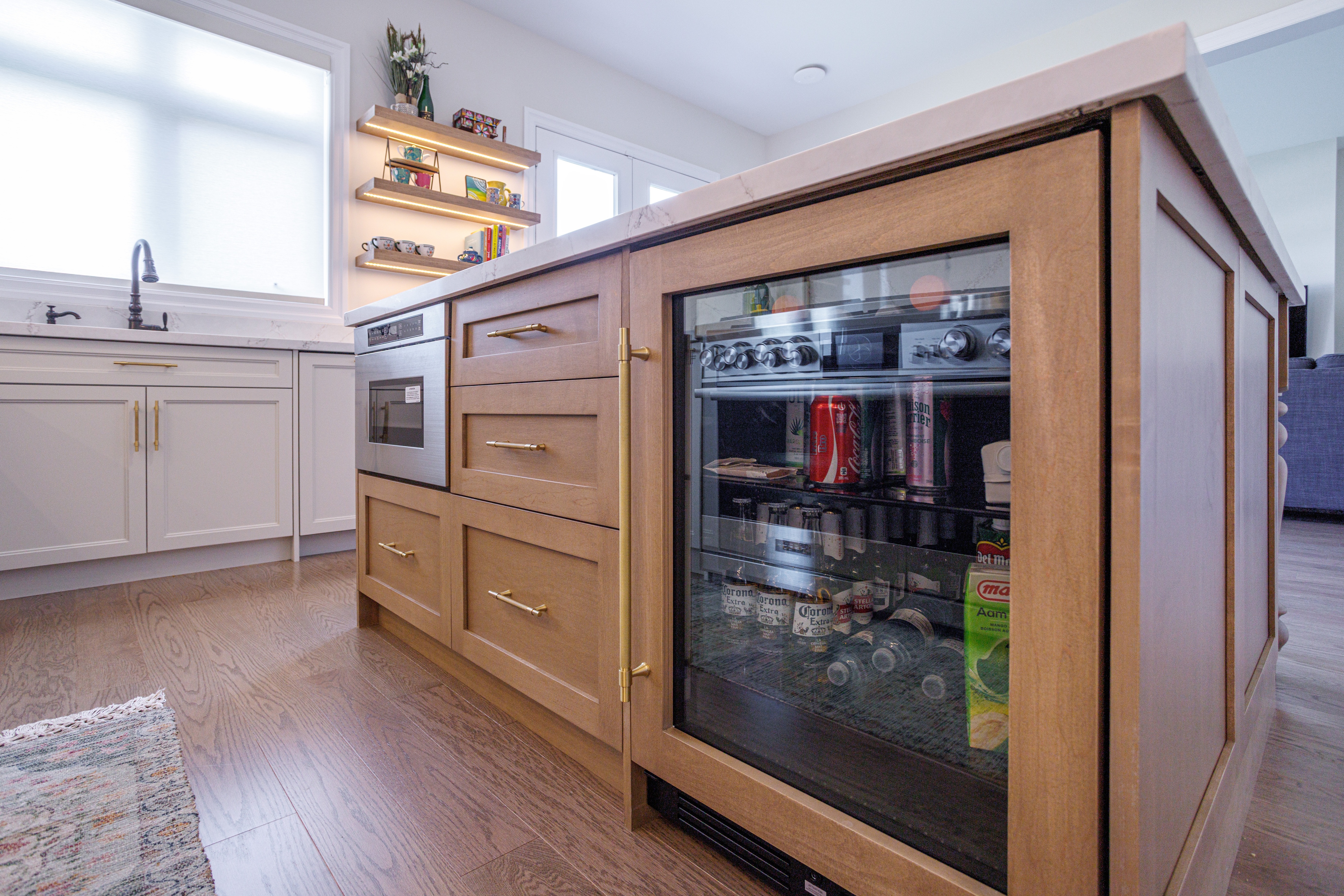 Kitchen island with wood cabinetry, brass handles, a glass‑front beverage fridge stocked with drinks, built‑in microwave, and marble countertop, with white cabinets and open shelving visible in the background.