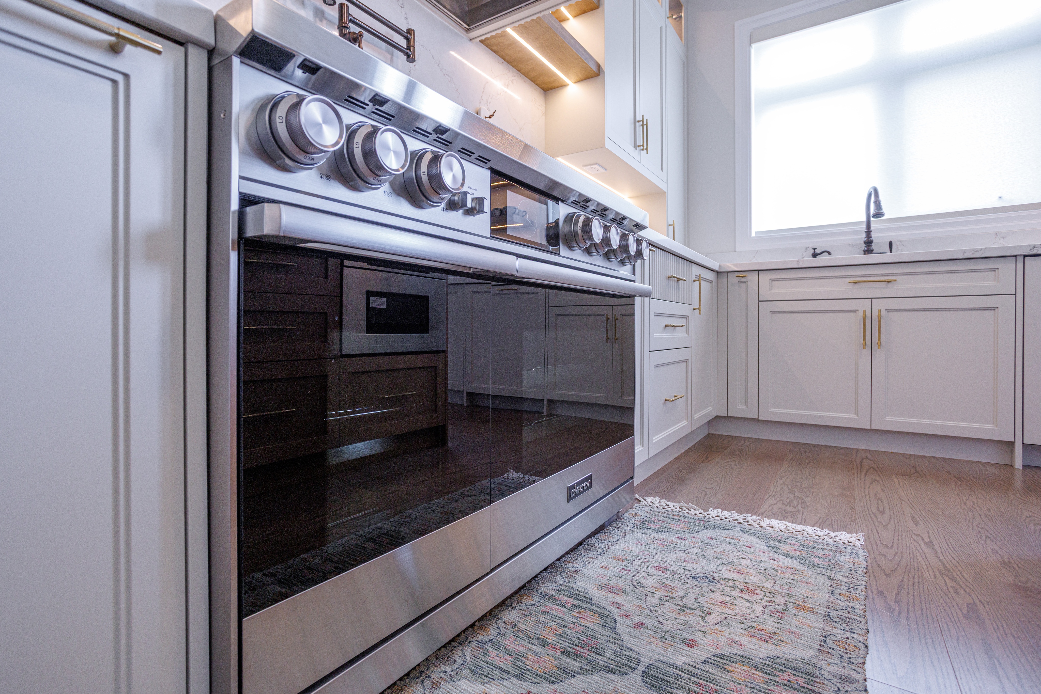 Stainless steel oven built into white cabinetry with gold handles, marble countertop above, patterned rug on wood flooring, and a sink beneath a large frosted window in a modern kitchen.